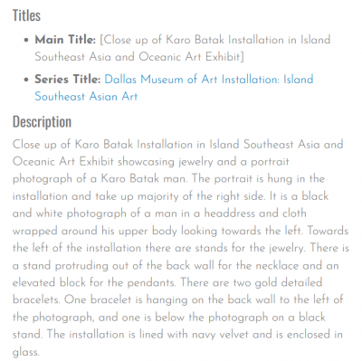 Close up of Karo Batak Installation in Island Southeast Asia and Oceanic Art Exhibit showcasing jewelry and a portrait photograph of a Karo Batak man. The portrait is hung in the installation and take up majority of the right side. It is a black and white photograph of a man in a headdress and cloth wrapped around his upper body looking towards the left. Towards the left of the installation there are stands for the jewelry. There is a stand protruding out of the back wall for the necklace and an elevated block for the pendants. There are two gold detailed bracelets. One bracelet is hanging on the back wall to the left of the photograph, and one is below the photograph on a black stand. The installation is lined with navy velvet and is enclosed in glass.